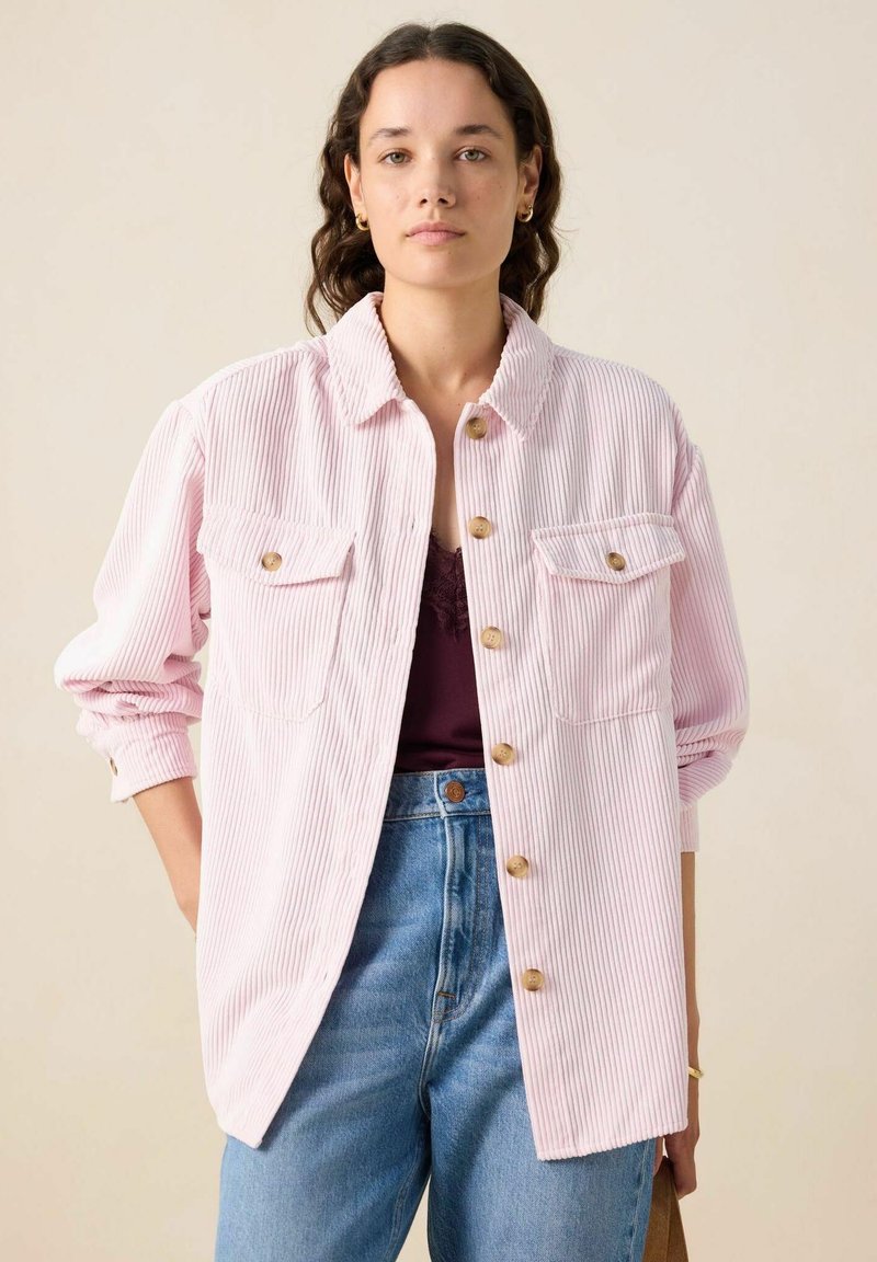 Roze corduroy oversized overhemdjack met knoopsluiting en borstzakken, gedragen over een donker topje, gecombineerd met blauwe high-waisted jeans.