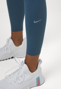 Gambe che indossano leggings Nike blu e scarpe Nike bianche con accenti turchesi e rosa su uno sfondo bianco.
