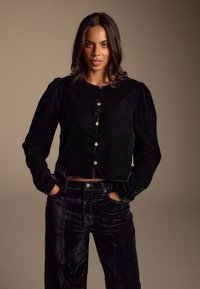 Zwarte cropped jas van fluweel met lange pofmouwen, voorzien van een ronde halslijn en gouden knopen. Gecombineerd met zwarte fluwelen broek met hoge taille.