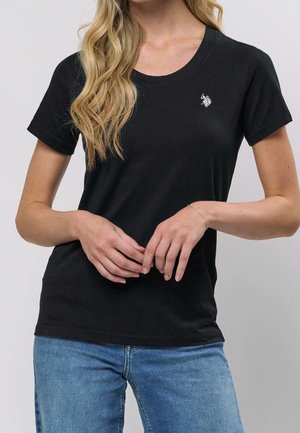 Femme blonde portant un t-shirt noir manches courtes avec un petit logo blanc sur la poitrine gauche et un jean bleu, mains jointes devant.