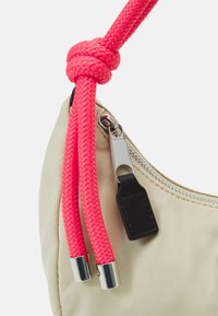 Beige väska med silverdragkedja och svart dragknapp, prydd med en rosa flätad rem som är knuten i en enkelknut med metalldelar.