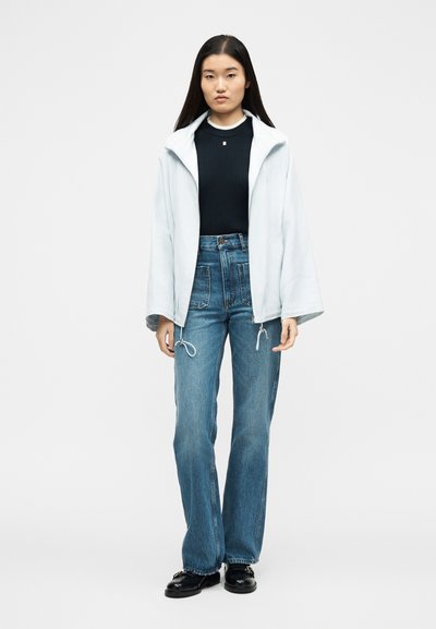 Νεαρή γυναίκα στέκεται φορώντας ελαφρύ μπουφάν πάνω από μαύρο πουλόβερ, τζιν με ψηλή μέση και μαύρα μοκασίνια, μπροστά σε λευκό φόντο.