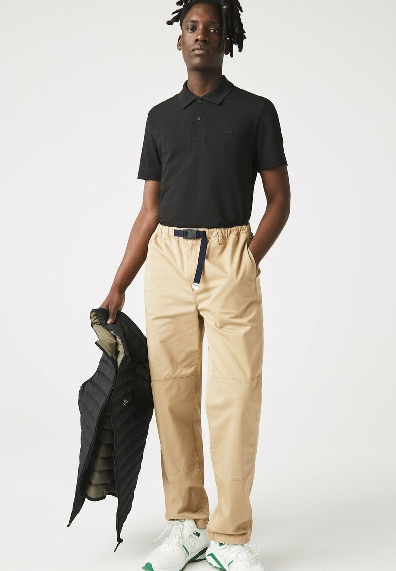 Polo noir, pantalon à taille élastique beige, gilet matelassé noir porté, et baskets blanches avec accents verts. Accent sur la fonctionnalité et le confort.