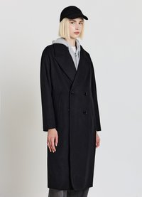 Cappotto doppiopetto nero con grandi rever, tasche laterali e tessuto strutturato; abbinato a una felpa con cappuccio grigia e un berretto nero.