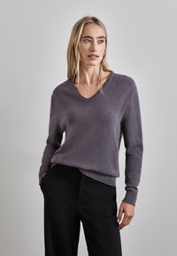 V-kauluspaita pehmeässä, hillityssä violetissa neuloksessa, jossa on pitkät hihat ja ribbaushelma. Yhdistetty mustiin housuihin, esitetty neutraalin taustan edessä.
