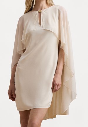 Vrouw draagt een crèmekleurige, knielange jurk met een vloeiende cape-achtige overlay en een kleine sleutelgathalslijn met een gouden gesp.