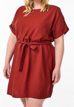 Woman wearing a short-sleeve, rust-colored dress with rolled cuffs and a tied belt at the waist, standing against a white background.