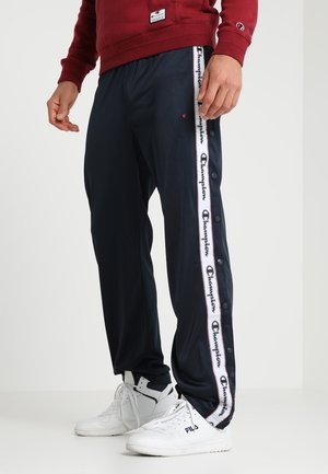 Person trägt schwarze Champion-Trainingshose mit seitlichen Druckknöpfen und weiße FILA-Sneaker, kombiniert mit einem roten Sweatshirt.