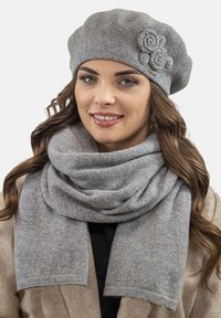 Grå uldberet med blomsterdetaljer og matchende grå uldscarf, der har en blød tekstur og vævet mønster, båret over en beige frakke.