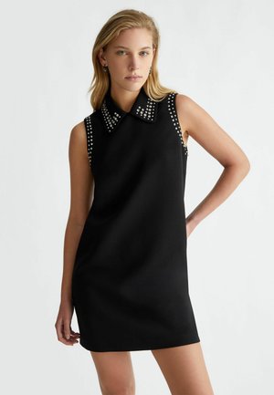 Abito nero senza maniche con una silhouette aderente, caratterizzato da un ampio colletto decorato con borchie metalliche lungo il colletto e le armature.
