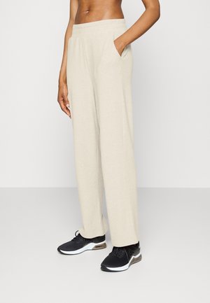 Personne portant un pantalon beige à jambes larges avec des poches et des chaussures de sport noires, debout devant un fond clair uni.