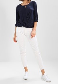 Kvinna klädd i en marinblå långärmad topp med rynkad framdel, vita slim-fit byxor och vita sneakers, stående mot en enkel ljus bakgrund.