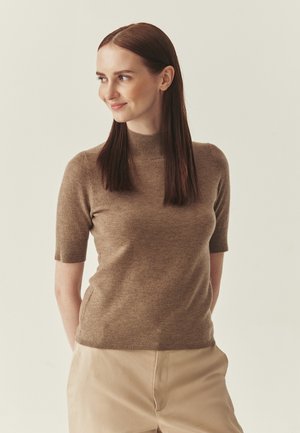 Młoda kobieta o prostych brązowych włosach, ubrana w beżowy krótki sweter z golfem i jasne spodnie, patrząca w lewą stronę, lekko się uśmiechająca.