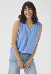 Blouse sans manches bleu clair en tissu texturé, avec décolleté en V et détail de bouton unique, associée à un jean en denim clair.