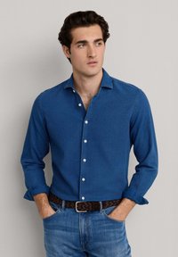 Mladý muž s tmavými vlasmi, oblečený v zapnutej modrej košeli a modrých džínsoch s hnedým pleteným opaskom, ruky v vreckách, pozerajúci sa bokom.