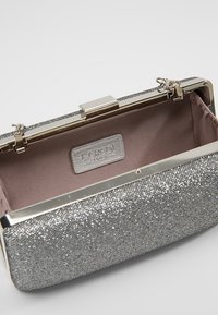 Pochette argentée scintillante avec un extérieur texturé, une quincaillerie métallique et un intérieur en tissu rose pâle. Fermeture à pression en haut avec détail logo.