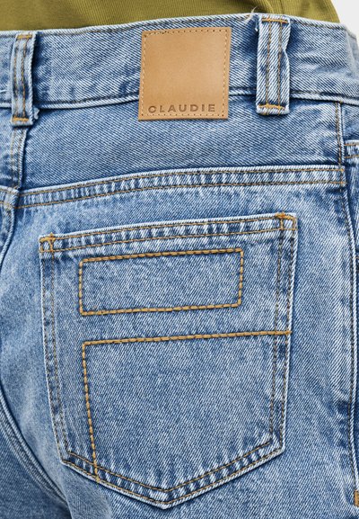 Ανοιχτό μπλε τζιν τζιν με ψηλή μέση, με δερμάτινο ταμπελάκι με την επιγραφή «CLAUDIE» και αντίθετες κίτρινες ραφές στις τσέπες.