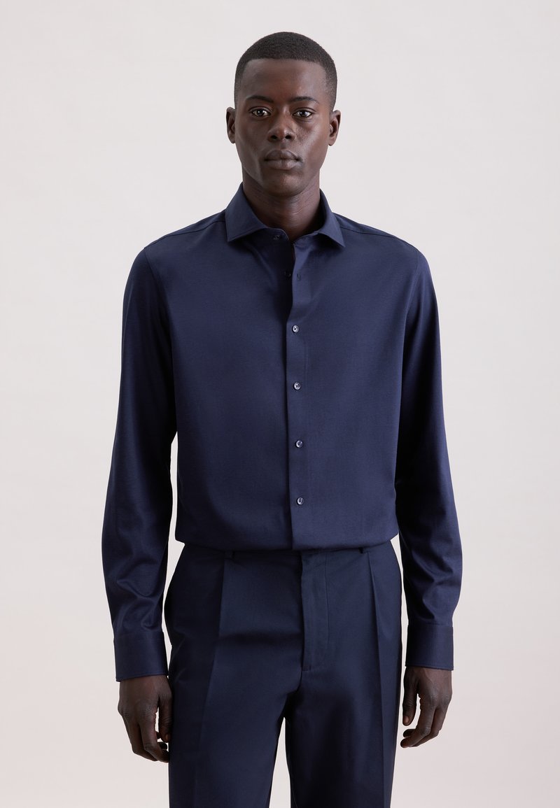 Homme portant une chemise bleu foncé à manches longues boutonnée et un pantalon bleu foncé assorti, debout devant un fond clair uni.