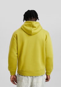 Sudadera amarilla con un corte relajado, capucha ajustable, puños acanalados y dobladillo. Textura suave con un diseño simple, vista desde la parte de atrás.