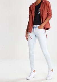 Lyseblå skinny jeans, en sort t-shirt med grafik, og en rustrød, let vævet hættetrøje. Hvide sneakers fuldender outfittet.
