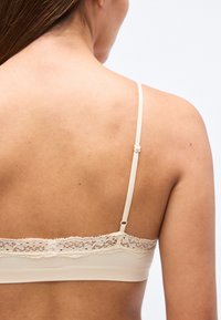 Μπεζ bralette με απαλό, ελαστικό ύφασμα, δαντέλα στο πάνω μέρος και ρυθμιζόμενες στενές τιράντες. Σχεδιασμός με λεία πλάτη και μινιμαλιστικό στυλ.