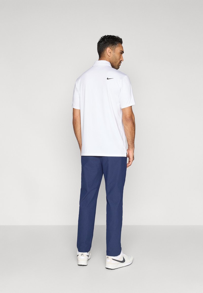 Polo blanc avec un logo Nike noir dans le dos, associé à un pantalon bleu marine et des chaussures blanches. Le polo a des manches courtes et un col.