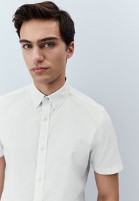 Chemise blanche à manches courtes avec col, en tissu lisse, elle présente un logo discret sur la poitrine gauche et une patte de boutonnage. Design simple.