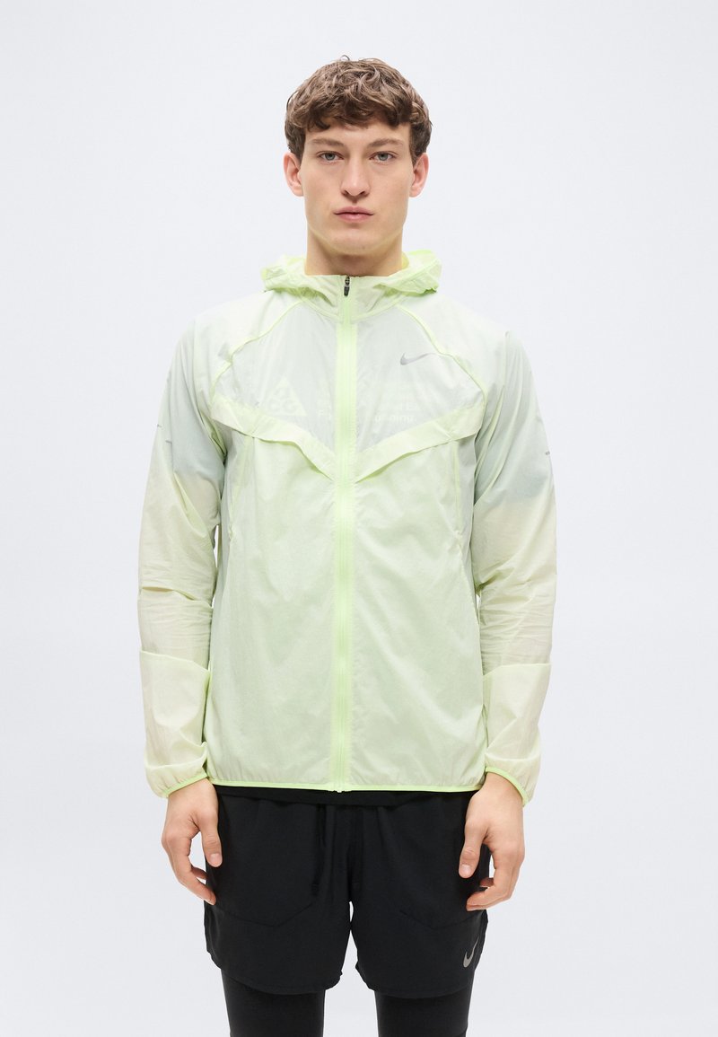 Ελαφρύ, ημιδιάφανο μπουφάν σε νέον πράσινο χρώμα με κουκούλα, κλείσιμο με φερμουάρ και αθλητική γραμμή, με αντανακλαστικές λεπτομέρειες και διακριτά τμήματα από διχτυωτό ύφασμα.