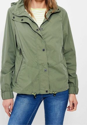 Femme portant une veste à capuche vert olive avec fermeture éclair et boutons, poignets élastiques et jean bleu, vue du cou jusqu'aux hanches.