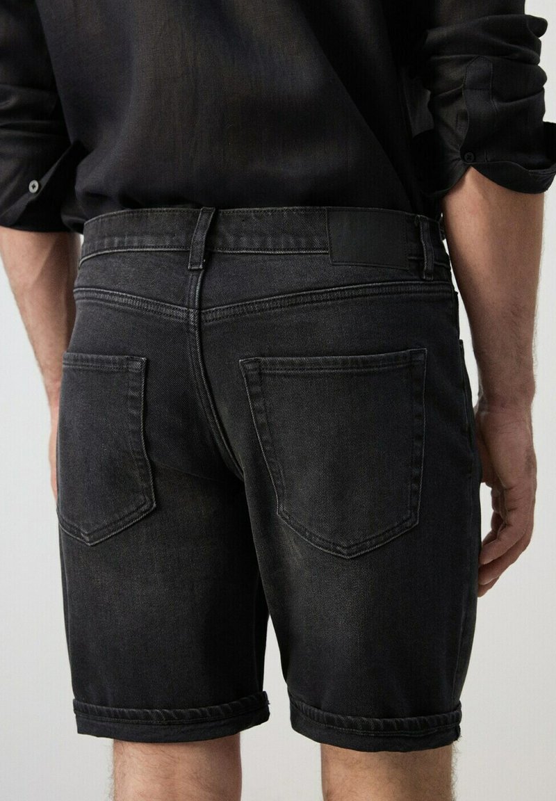 Homme portant un short en denim noir avec des poches arrière et une chemise noire à manches longues retroussées, vu de dos.