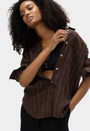 Camicia marrone a righe con bottoni, vestibilità rilassata, con una leggera lucentezza, maniche arrotolate e dettagli a righe bianche.