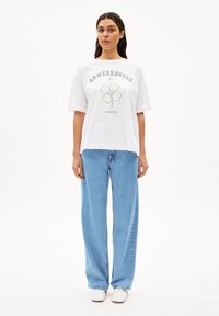 Witte katoenen t-shirt met een groen vierbladig klavertje ontwerp en de tekst "ARMEDANGELS" erboven. Gecombineerd met lichtblauwe wijde jeans en witte schoenen.