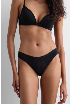 Femme portant un soutien-gorge noir uni assorti et un slip bikini taille haute, debout devant un fond gris clair neutre.