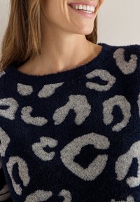 Granatowy sweter o miękkiej, puszystej fakturze z dużymi beżowymi plamami w panterkę. Okrągły dekolt i widoczne ramiona.