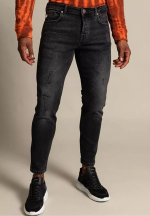 Homme portant un jean slim noir déchiré, des baskets noires avec des semelles blanches et une chemise orange à manches longues, debout sur un fond neutre.