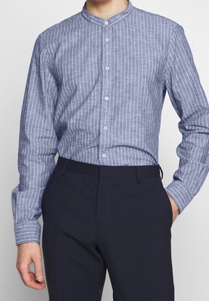 Mand iført en blå langærmet skjorte med hvide lodrette striber og sorte bukser, stående med den ene hånd i lommen.