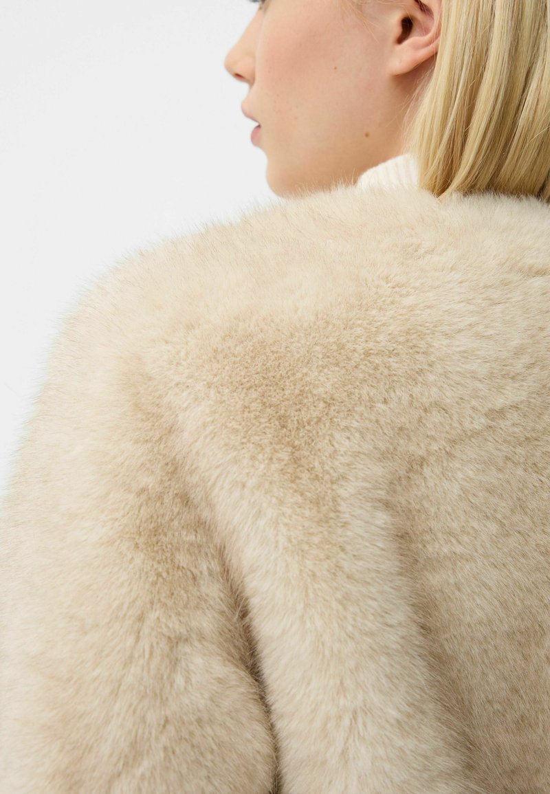 Cappotto in pelliccia sintetica beige con una superficie morbida e texture, caratterizzato da un ampio colletto e una leggera lucentezza. Cuciture visibili sulle spalle.