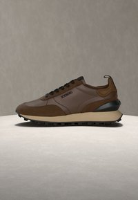 Baskets en cuir marron et suède avec lacets noirs, semelle beige, semelle extérieure noire et logo ICENRG sur le côté, sur une surface claire et un fond gris.