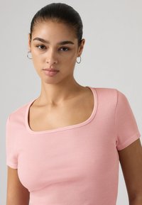 Νεαρή γυναίκα με σκούρα μαλλιά σε κότσο, που φοράει κρίκους στα αυτιά και ροζ ριγωτό μπλουζάκι με κοντά μανίκια και τετράγωνη λαιμόκοψη, κοιτάζοντας μπροστά.