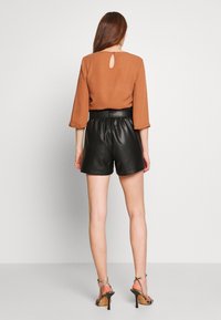 Hnědá blůza s kulatým klíčovým otvorem na zadní straně a tříčtvrtečními rukávy, spárovaná s černými šortkami z umělé kůže se zvýšeným pasem a páskovými podpatky.