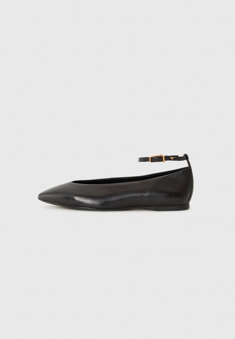 Chaussure plate en cuir noir à bout pointu avec une fine bride à la cheville et une boucle dorée sur un fond gris clair uni.