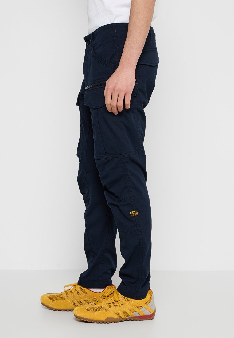 Person, der marineblaue Cargo-Hosen mit mehreren Taschen und gelben Sportschuhen trägt, steht vor einem weißen Hintergrund.