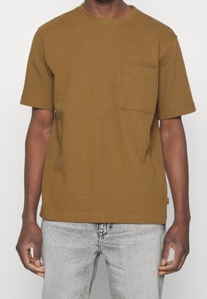 Brun tekstureret t-shirt med kort ærme, rund hals og lomme på venstre bryst, parret med lysegrå jeans.