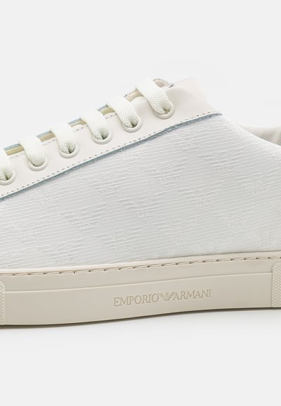 White sneaker with textured fabric, flat laces, and a pale blue accent near the eyelets. Features engraved logo on the sole.