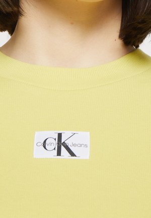 Nahaufnahme einer Person, die ein gelb geripptes Shirt trägt, auf dem sich mittig unterhalb des Halsausschnitts ein rechteckiges weißes Logo von Calvin Klein Jeans befindet.