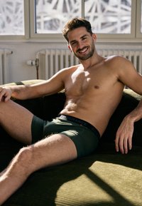 Boxer aderenti di colore verde scuro con un bordo nero "ENDURANCE", design fitted e texture visibile. Il soggetto è seduto su un divano verde.