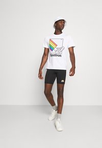 Hvid Reebok t-shirt med farverige regnbue grafik og logo, kombineret med sorte shorts og hvide sneakers. Lys grå baggrund.