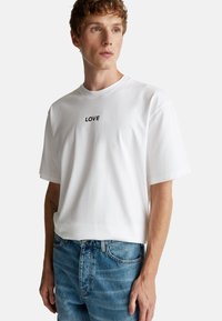 Camiseta de algodón blanca con cuello redondo, que presenta la palabra "AMOR" en texto negro en el pecho. Combinada con jeans de mezclilla azules.