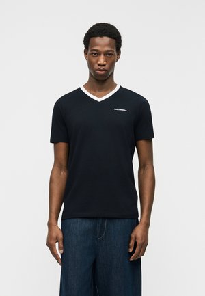 Mężczyzna ubrany w czarną koszulkę z krótkim rękawem i dekoltem w serek z białym obszyciem oraz małym białym napisem „KARL LAGERFELD”, w zestawieniu z ciemnoniebieskimi dżinsami.