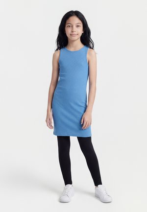 Dievča stojace s dlhými tmavými vlasmi, oblečené v modrých rebrovaných šatách bez rukávov, čiernych legínach a bielych teniskách na bielom pozadí.
